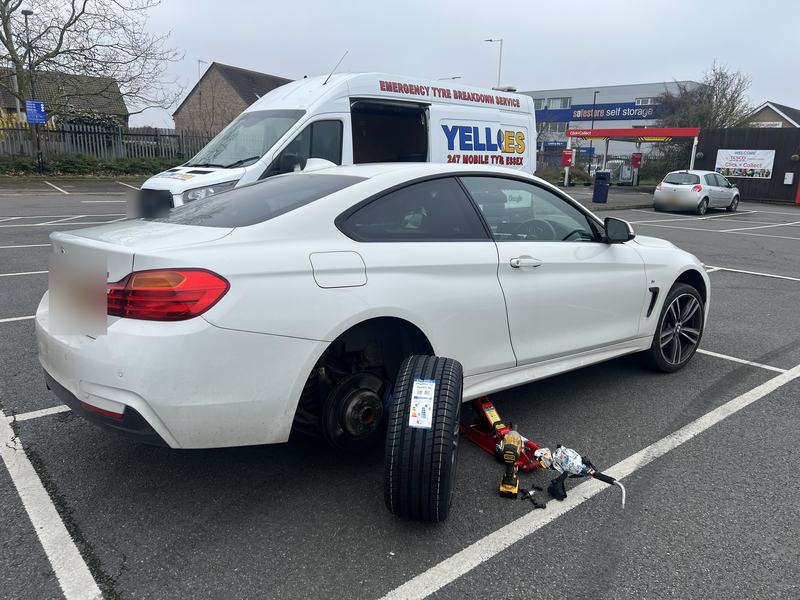 White BMW 4 Series Gran Coupe with a rear wheel off in a supermarket car park, new tyre with its EU label ready to fit