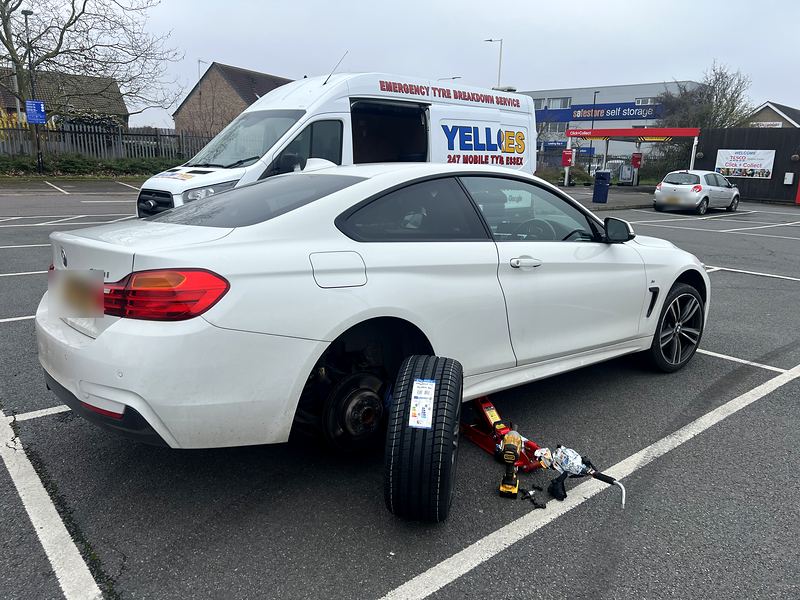 New tyre with EU label beside a BMW coupe on a jack in a supermarket car park, Yellow Tyres van behind