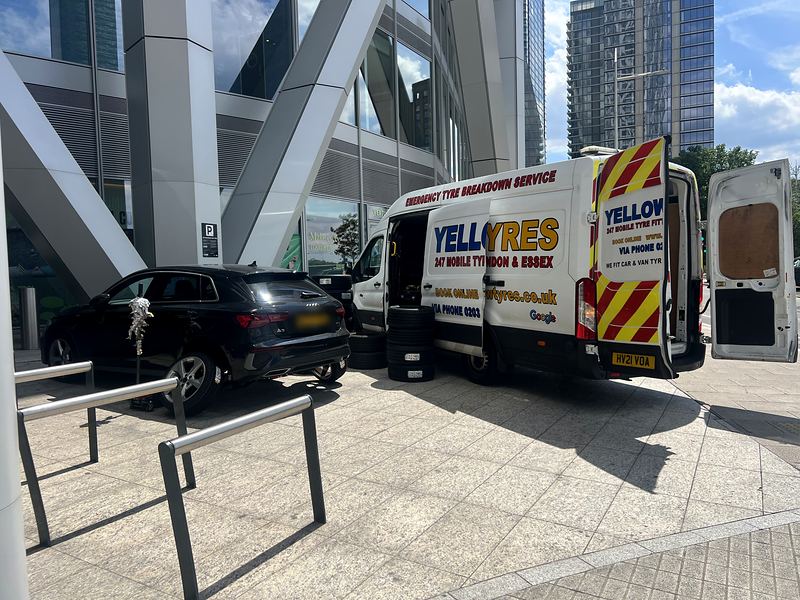 Yellow Tyres van with rear doors open beside an Audi on a jack outside glass office towers in East London