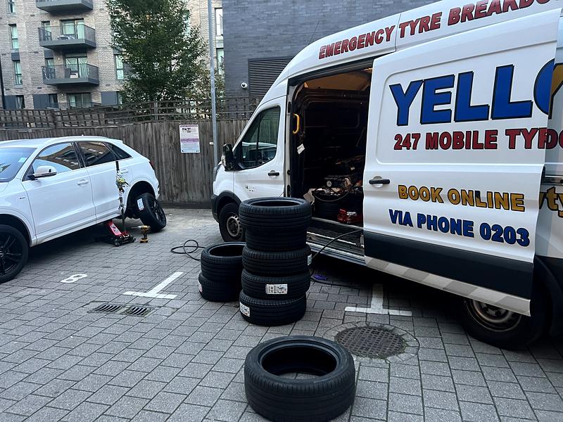 Yellow Tyres van with side door open and new tyres stacked beside an SUV on a jack in a courtyard car park