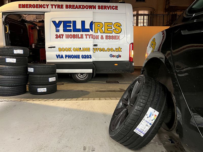New tyre with its EU label still on, leaning against a car, Yellow Tyres van and tyre stock behind