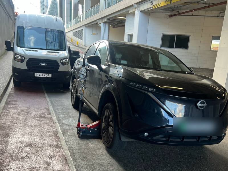 Black Nissan Ariya electric SUV on a trolley jack under a building canopy, Yellow Tyres van parked behind